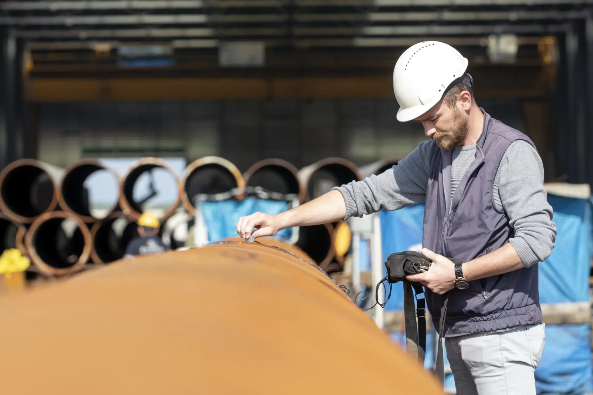 An inspector in a hard hat checking a weld seam on large-diameter steel pipe in a fabrication yard