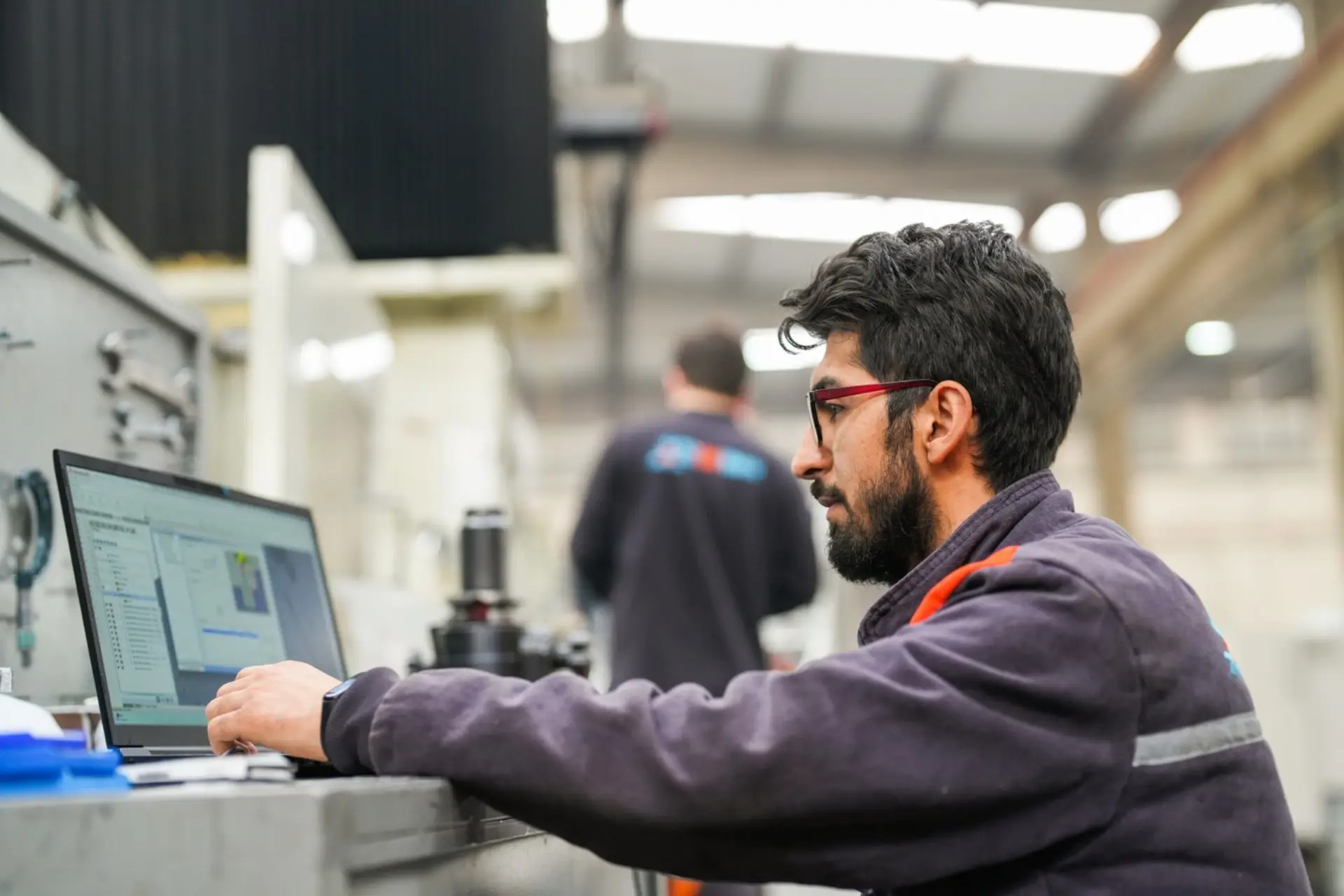 An engineer on a shop floor reviewing machining software on a laptop beside a milling centre