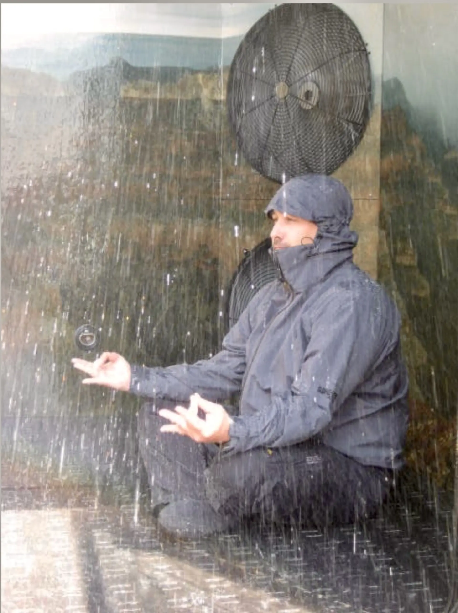 Inside the rain room: a person in a hooded waterproof jacket sits cross-legged under steady falling rain, hands open and palms up, with a large industrial fan mounted on the wall behind them.